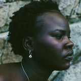 A close-up side profile of a woman with short dark curly hair, wearing a delicate silver necklace with a small red and white gemstone pendant and a matching silver hoop earring, set against a textured gray stone wall.