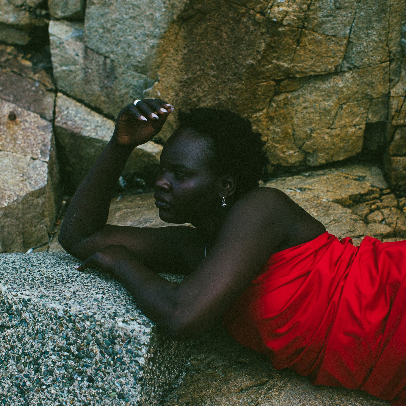 A woman reclining on large stone blocks outdoors, wearing a bright red draped garment. The background consists of natural rock formations.