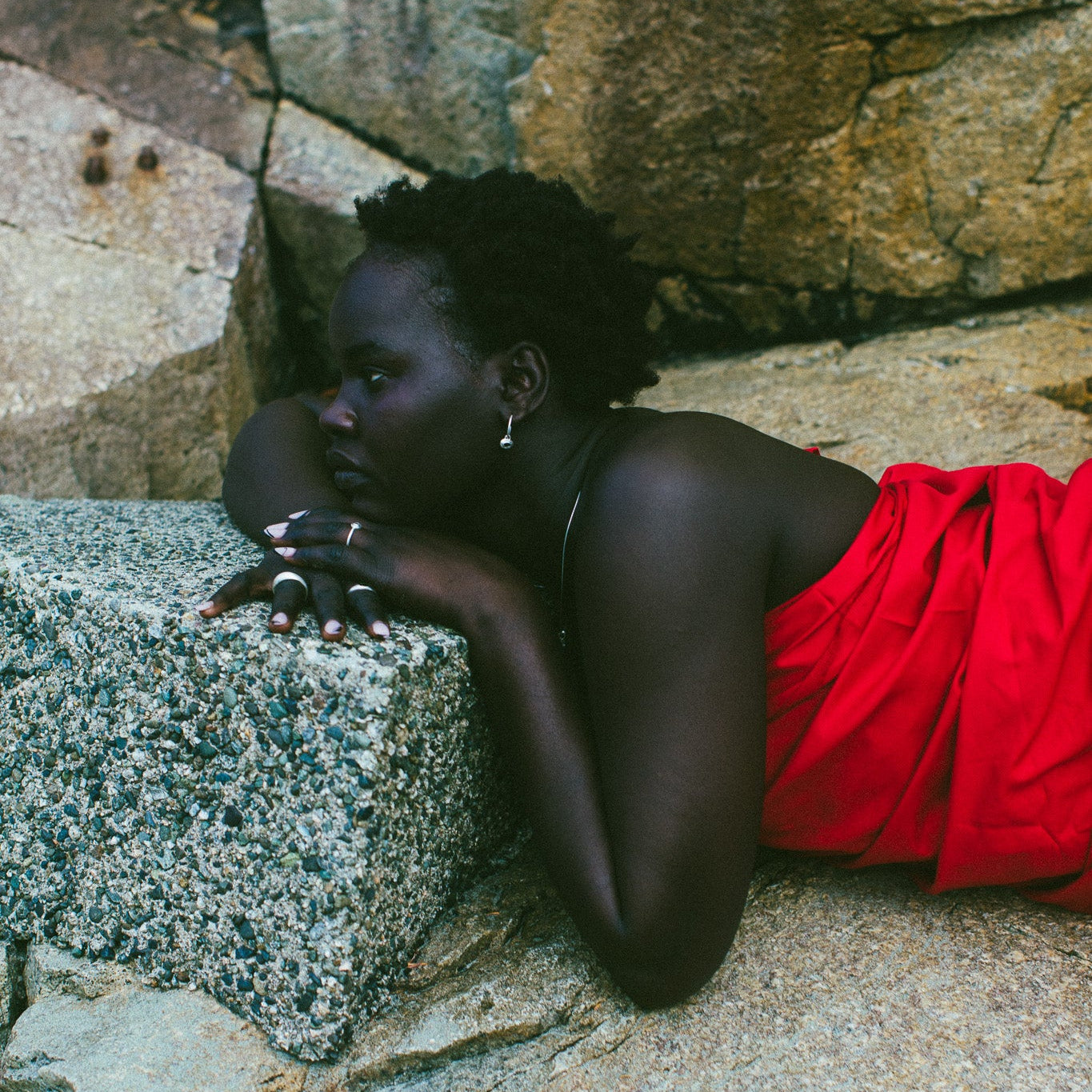 A woman with short dark hair lies on large gray rocks, wearing a bright red draped garment and small hoop earrings.