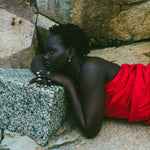 A woman with short dark hair lies on large gray rocks, wearing a bright red draped garment and small hoop earrings.