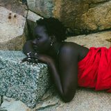 A woman with short dark hair lies on large gray rocks, wearing a bright red draped garment and small hoop earrings.