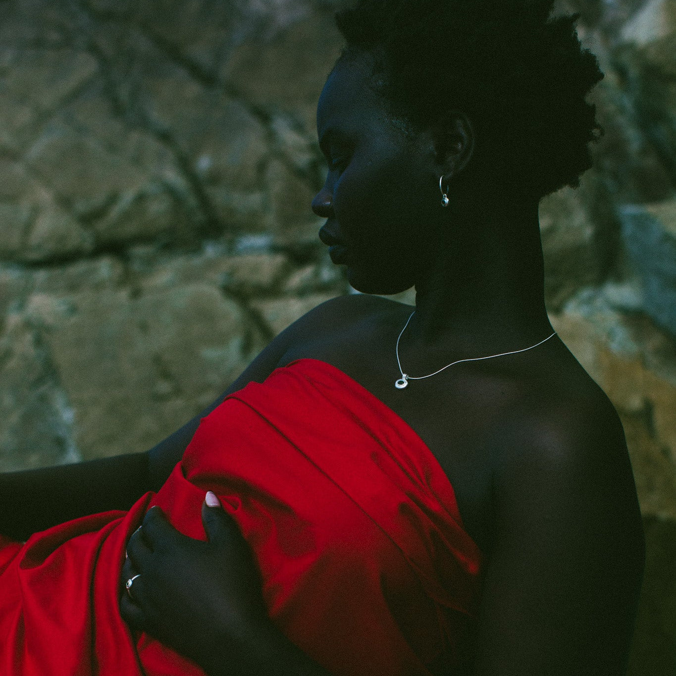 A woman in profile wearing a bright red draped dress or wrap, posed against a dark stone wall. She is wearing a delicate silver necklace and a ring.