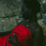 A woman in profile wearing a bright red draped dress or wrap, posed against a dark stone wall. She is wearing a delicate silver necklace and a ring.