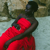 A woman seated outdoors on a metal chair against a rocky, sandy background, wearing a bright red strapless draped dress.