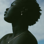 A close-up side profile of a person outdoors against a cloudy blue sky, wearing a delicate silver hoop earring and a thin silver necklace.