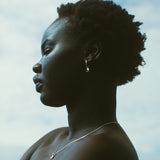 A side-profile portrait of a woman with short curly hair, wearing a red strapless top, a delicate silver necklace, and a small silver hoop earring against a cloudy sky background.