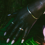 A close-up of a hand resting on bright green fern leaves outdoors. The hand is adorned with silver-toned jewelry, including a bangle bracelet and rings. One ring with an orange oval gemstone.