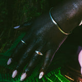 A close-up of a hand resting on bright green fern leaves outdoors. The hand is adorned with silver-toned jewelry, including a bangle bracelet and rings. One ring with an orange oval gemstone.
