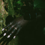 A close-up of a person's hand with long pale pink nails and several rings, resting against a mossy tree trunk in a dark outdoor setting with foliage.