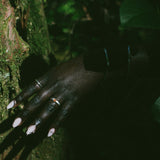 A close-up of a person's hand with long pale pink nails and several rings, resting against a mossy tree trunk in a dark outdoor setting with foliage.