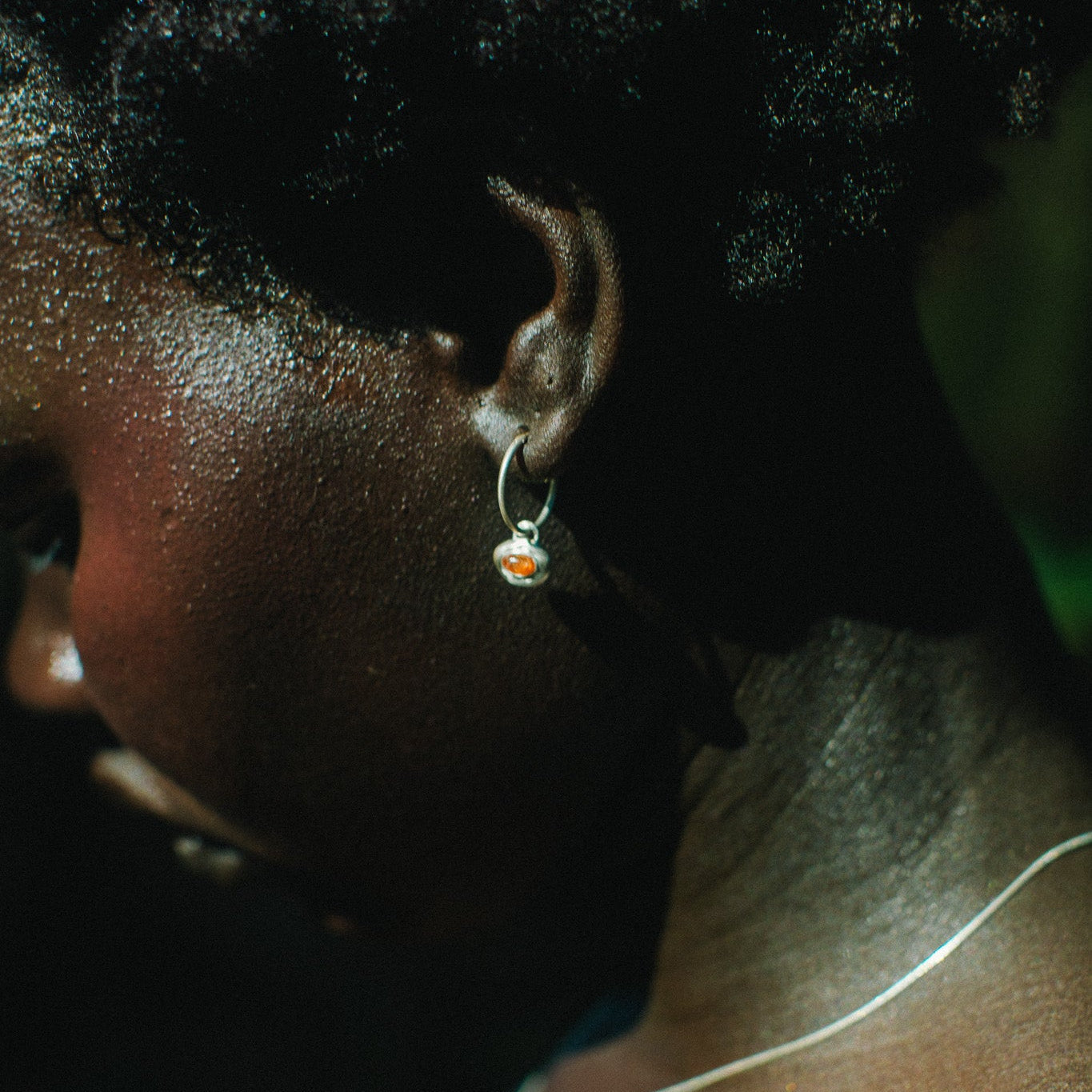 Close-up side profile of a person wearing a small silver hoop earring with a tiny orange and white stone, along with a thin silver necklace.