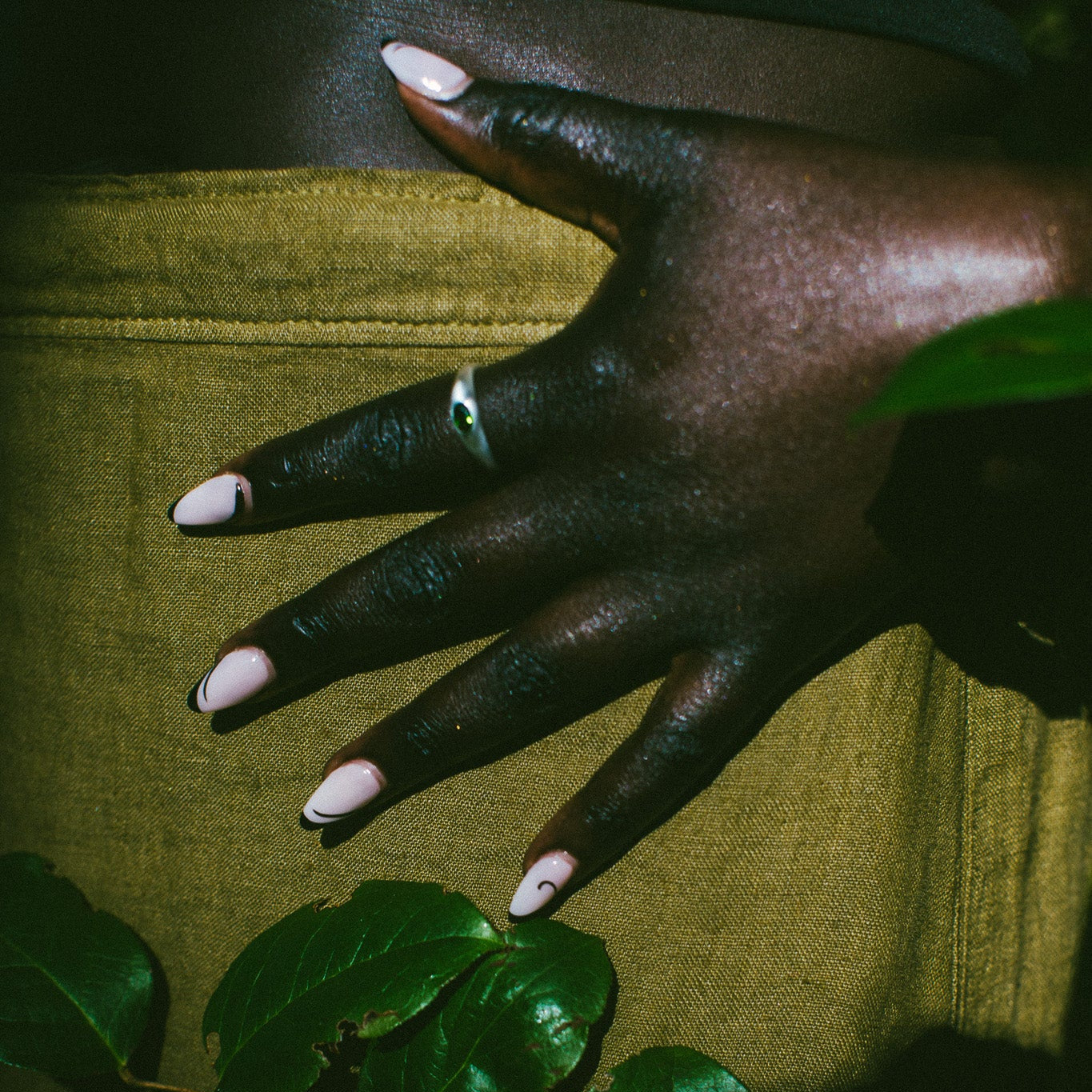 A close-up of a hand with long, pale pink manicured nails resting on a textured olive green fabric surface, with dark green leaves in the foreground and a silver ring on one finger.
