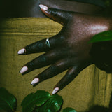 A close-up of a hand with long, pale pink manicured nails resting on a textured olive green fabric surface, with dark green leaves in the foreground and a silver ring on one finger.