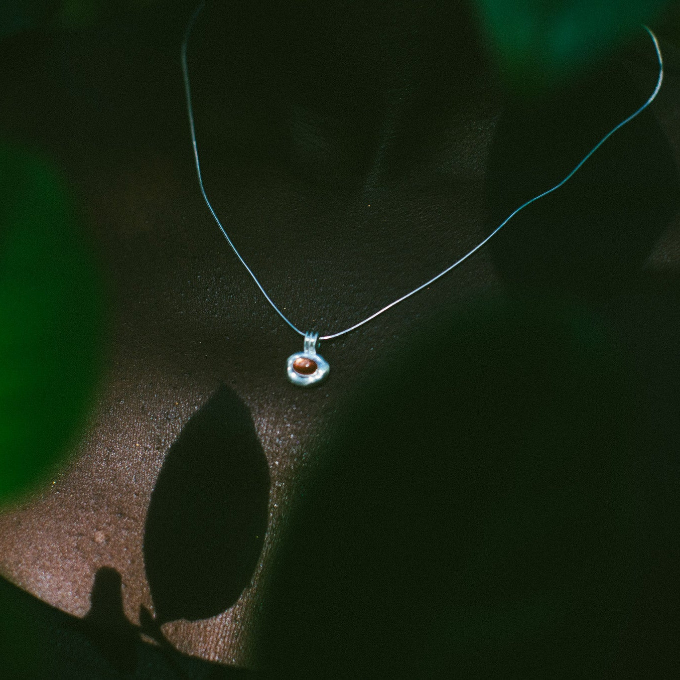 A delicate silver necklace with a small oval pendant featuring an orange stone design, photographed in dramatic, low-key lighting with dark green leaves in the foreground.