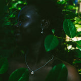 A woman in profile wearing a delicate silver necklace and earring, framed by lush green leaves in a dark, moody outdoor setting.