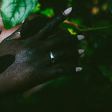 A close-up of a hand wearing a silver band ring, set against a dark background with green leaves.