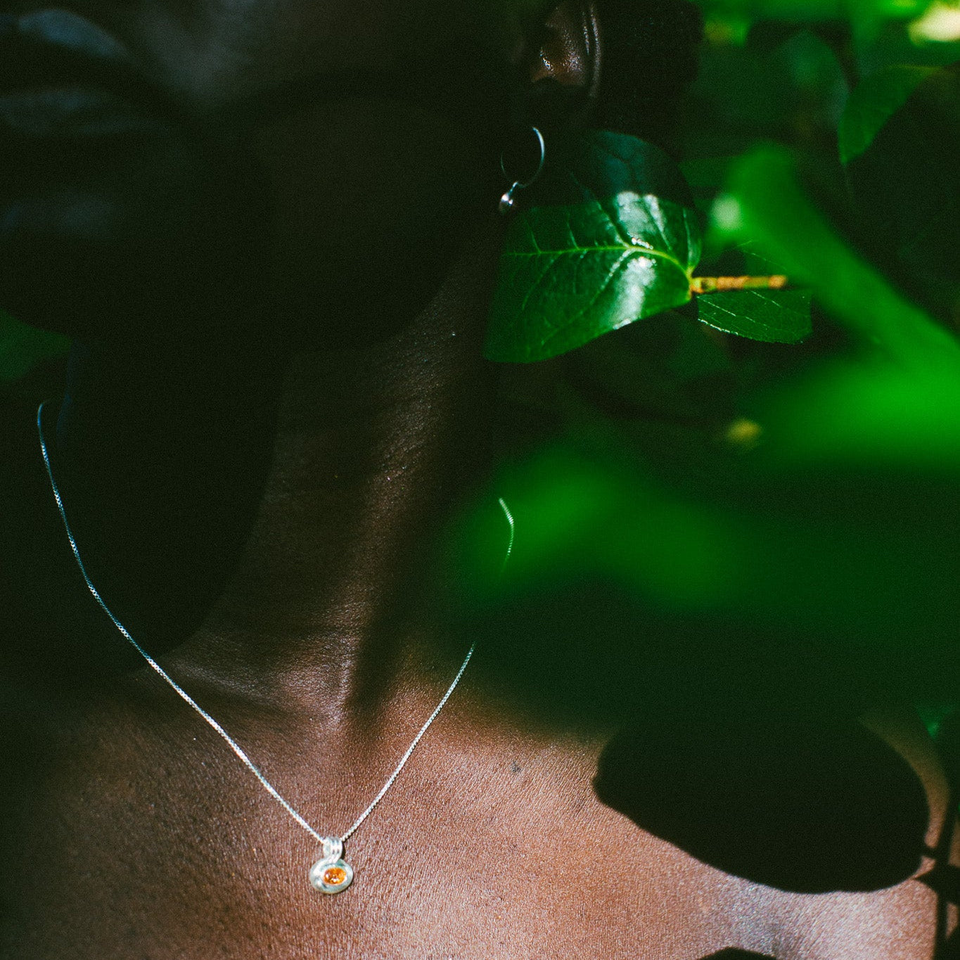 A close-up lifestyle shot of a model wearing a delicate silver chain necklace with a small round pendant, set against a dark background with dramatic green leaf shadows.