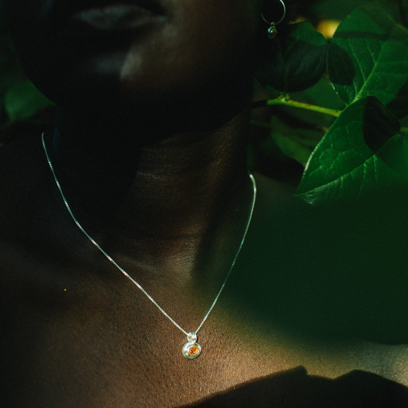 A close-up lifestyle shot of a woman wearing a delicate silver pendant necklace with a small orange and white stone, set against a dark background with green foliage.