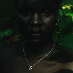 A close-up portrait of a person outdoors among dark green leaves, wearing a delicate silver necklace with a small circular pendant and hoop earrings.