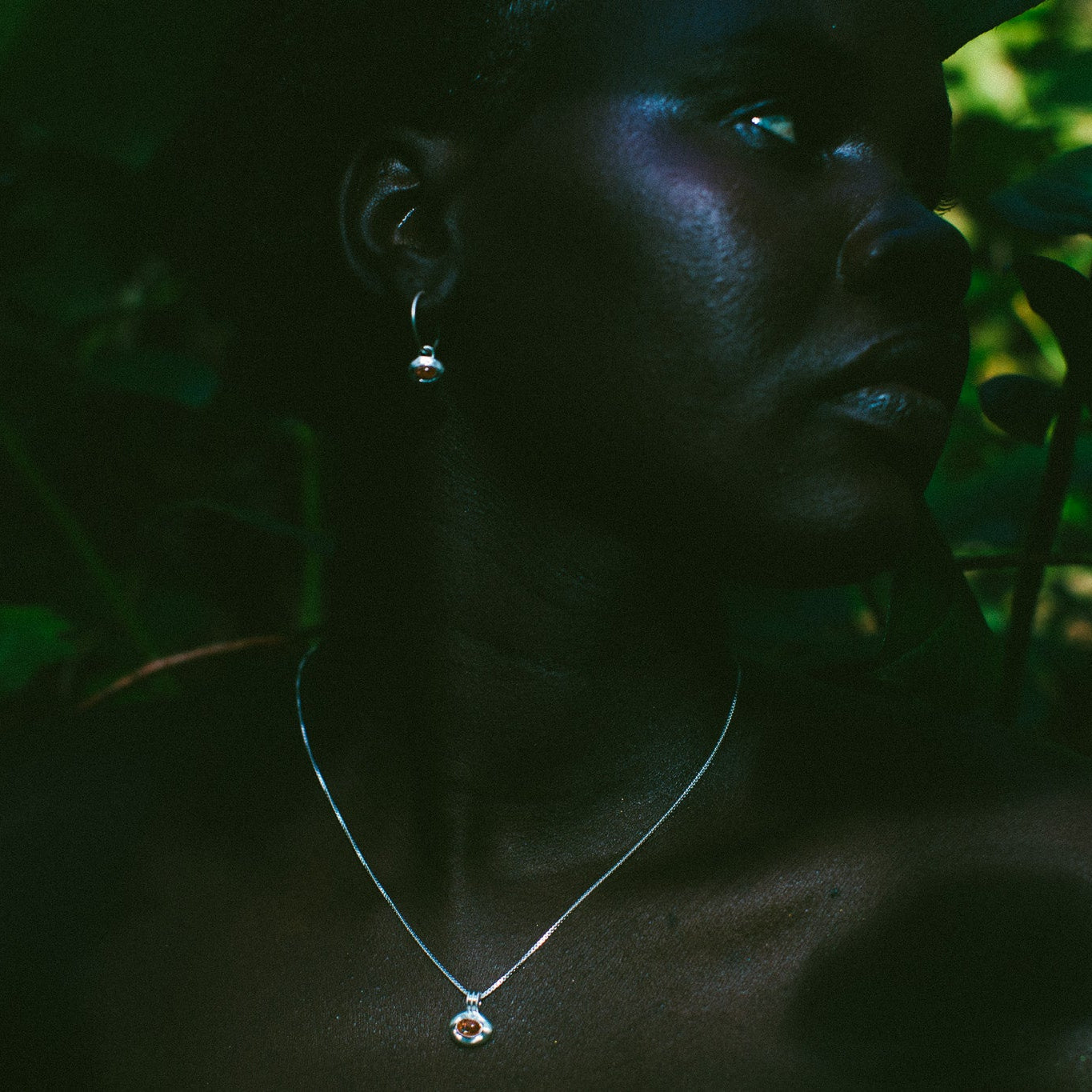 A close-up portrait of a woman in profile wearing a delicate silver pendant necklace and matching drop earring, set against a dark, leafy outdoor background.