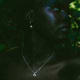 A close-up portrait of a woman in profile wearing a delicate silver pendant necklace and matching drop earring, set against a dark, leafy outdoor background.
