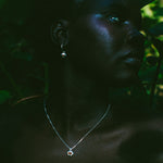 A close-up portrait of a woman in profile wearing a delicate silver pendant necklace and matching drop earring, set against a dark, leafy outdoor background.