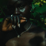 A close-up lifestyle shot of a person outdoors in a lush green environment, wearing a delicate silver necklace with a small clear pendant and a silver ring.