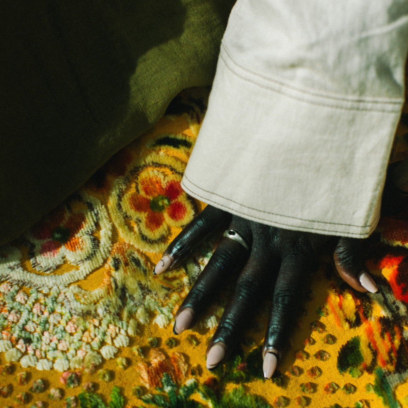 A close-up of a hand wearing a silver ring with an oval, green gemstone resting on a vibrant, multicolored floral patterned rug, with a light-colored denim cuff visible at the wrist.