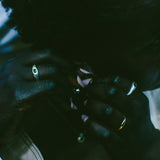 A close-up lifestyle shot of a person wearing multiple rings, including a prominent green stone ring on one finger, in low, moody, dark teal-toned lighting.