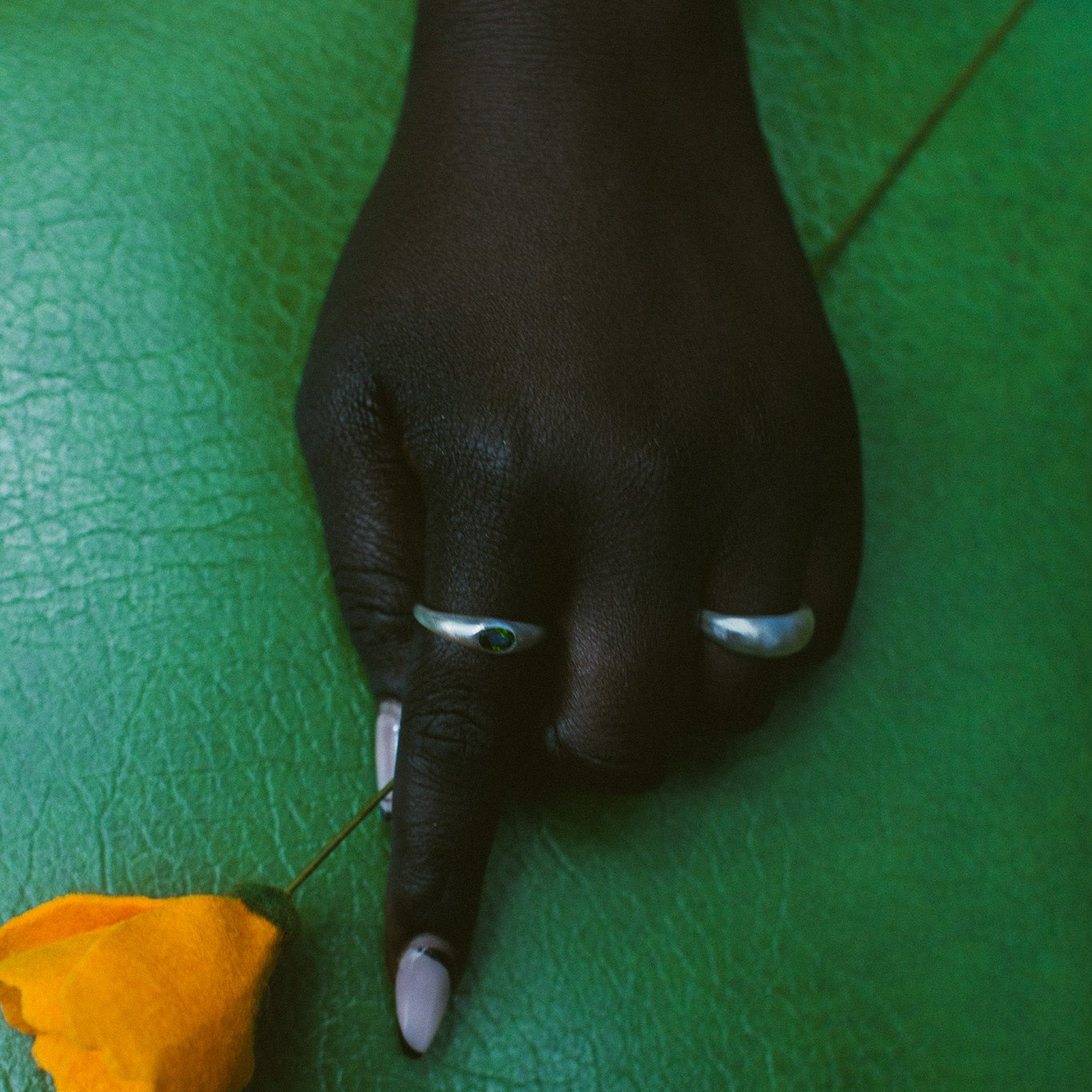 A close-up of a hand resting on a textured green surface, wearing two silver rings, with a small yellow flower nearby.