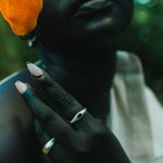 A close-up lifestyle shot of a person wearing two silver rings on their fingers, holding a bright orange flower near their face. The background is a dark, blurred outdoor setting with green foliage.