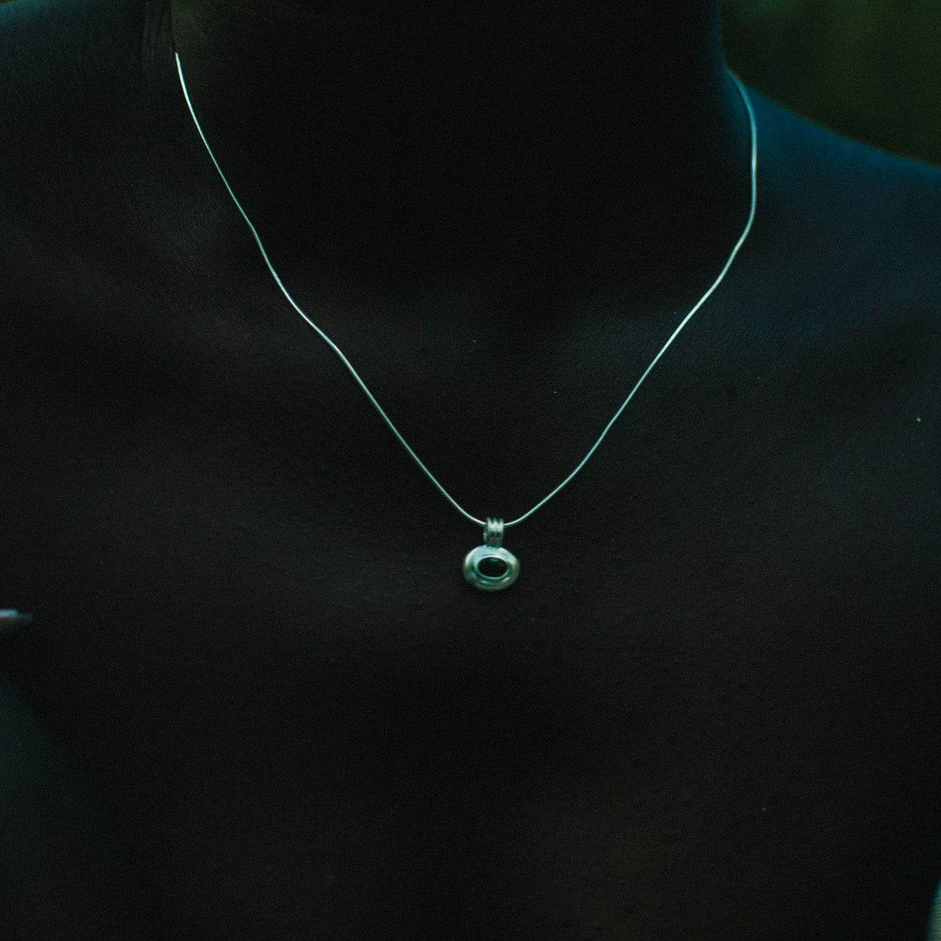 A close-up of a person wearing a delicate silver chain necklace with a small circular pendant featuring a green stone.
