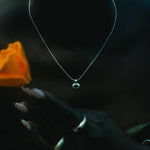 A close-up lifestyle shot of a person wearing a delicate silver chain necklace with a small round pendant, holding a vibrant orange rose. 