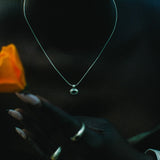 A close-up lifestyle shot of a person wearing a delicate silver chain necklace with a small round pendant, holding a vibrant orange rose. 