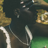 A close-up lifestyle shot of a woman with short black hair, wearing a silver necklace and earring, a white lace-trimmed top, and rings, resting her head on a green pillow.