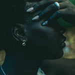Close-up side profile of a person with dark skin wearing silver drop earrings with manicured nails and rings visible against a dark, moody, forested background.