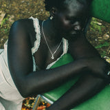 A woman with short dark hair sits on a green chair outdoors, wearing a white lace-trimmed camisole, and a silver necklace. A colorful patterned rug is visible beneath her.