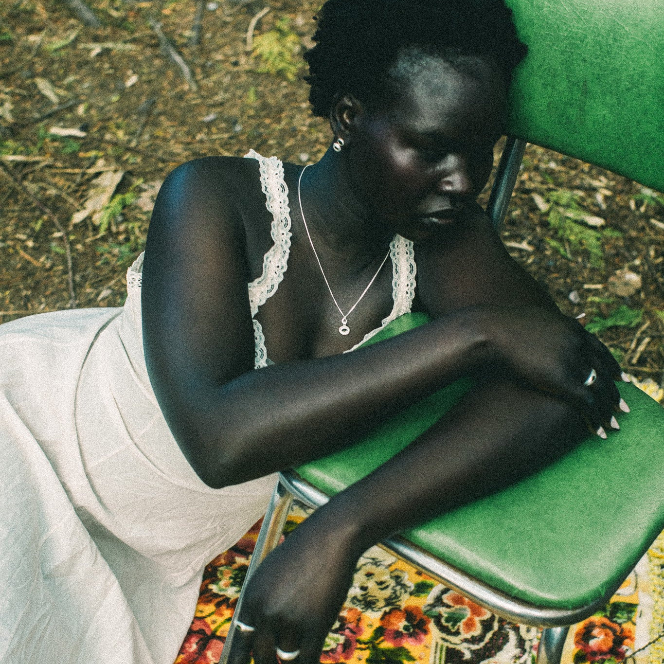 A woman sits slouching on a green metal chair outdoors, wearing a white sleeveless dress with lace trim and a silver necklace. She is resting on a colorful floral-patterned rug with forest floor foliage visible in the background.