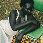 A woman sits slouching on a green metal chair outdoors, wearing a white sleeveless dress with lace trim and a silver necklace. She is resting on a colorful floral-patterned rug with forest floor foliage visible in the background.