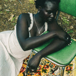 A woman sits slouching on a green metal chair outdoors, wearing a white sleeveless dress with lace trim and a silver necklace. She is resting on a colorful floral-patterned rug with forest floor foliage visible in the background.
