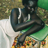 A woman sits slouching on a green metal chair outdoors, wearing a white sleeveless dress with lace trim and a silver necklace. She is resting on a colorful floral-patterned rug with forest floor foliage visible in the background.