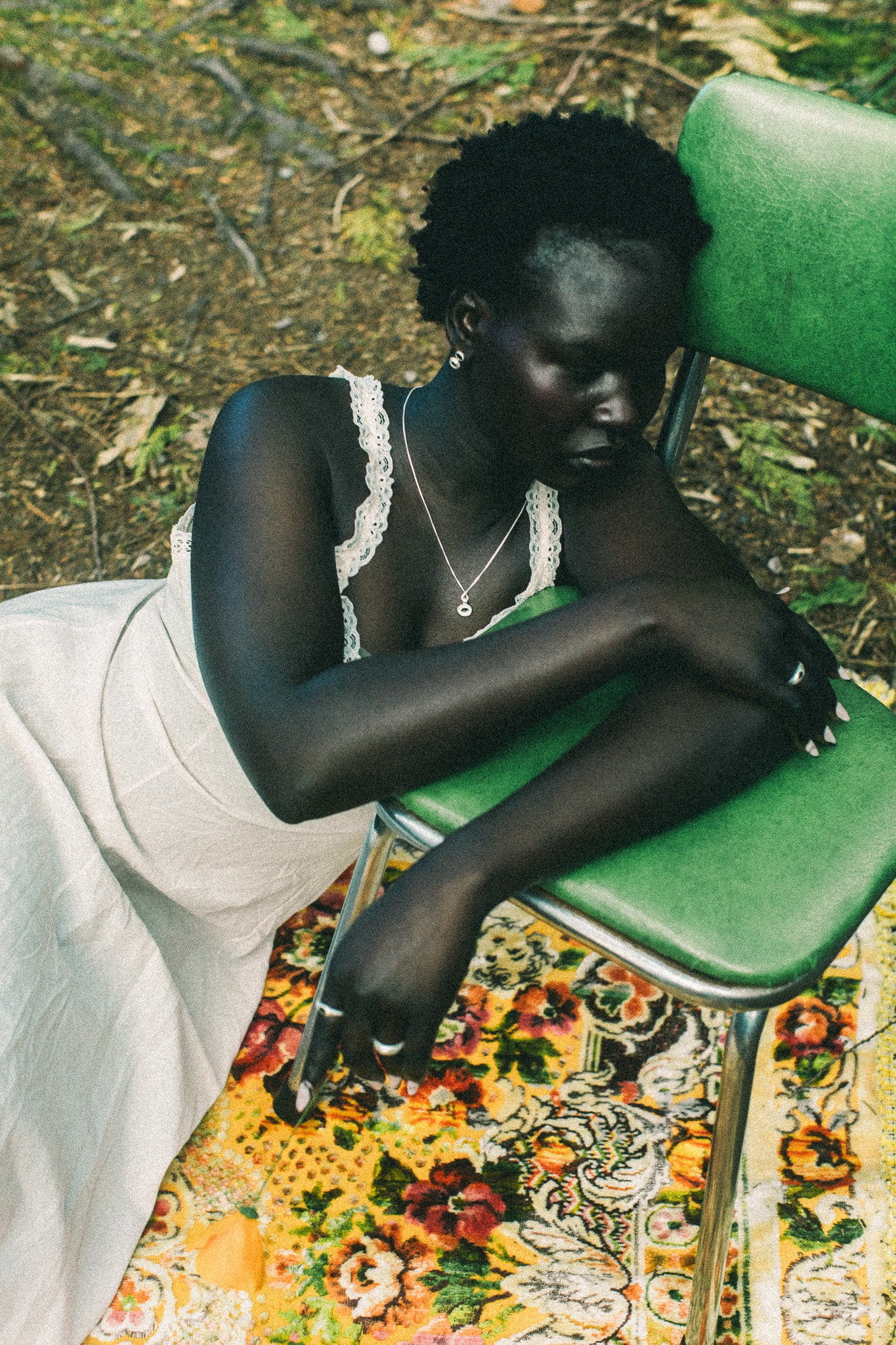 A woman sits slouching on a green metal chair outdoors, wearing a white sleeveless dress with lace trim and a silver necklace. She is resting on a colorful floral-patterned rug with forest floor foliage visible in the background.