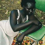 A woman sits slouching on a green metal chair outdoors, wearing a white sleeveless dress with lace trim and a silver necklace. She is resting on a colorful floral-patterned rug with forest floor foliage visible in the background.