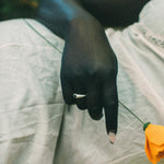 A close-up lifestyle shot of a person's hand resting on their lap, wearing a silver ring, with a small yellow flower held near their finger. They are dressed in a white skirt, set against a soft-focus outdoor background.