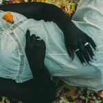 A person lying on a floral blanket, wearing a white lace-trimmed top and light gray skirt, with a single orange flower held near their chest. they are wearing rings.