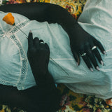 A person lying on a floral blanket, wearing a white lace-trimmed top and light gray skirt, with a single orange flower held near their chest. they are wearing rings.