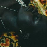 A close-up lifestyle shot of a person lying down, wearing a delicate silver stud dangle earring and a thin silver necklace, resting on a patterned yellow and red rug.