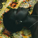A close-up lifestyle shot of a person lying down on a colourful floral patterned rug, wearing a delicate silver necklace and a small silver stud drop earring.