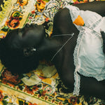 A person lying on their back on a colorful floral patterned blanket, wearing a white lace-trimmed top and a small orange flower tucked into the neckline.