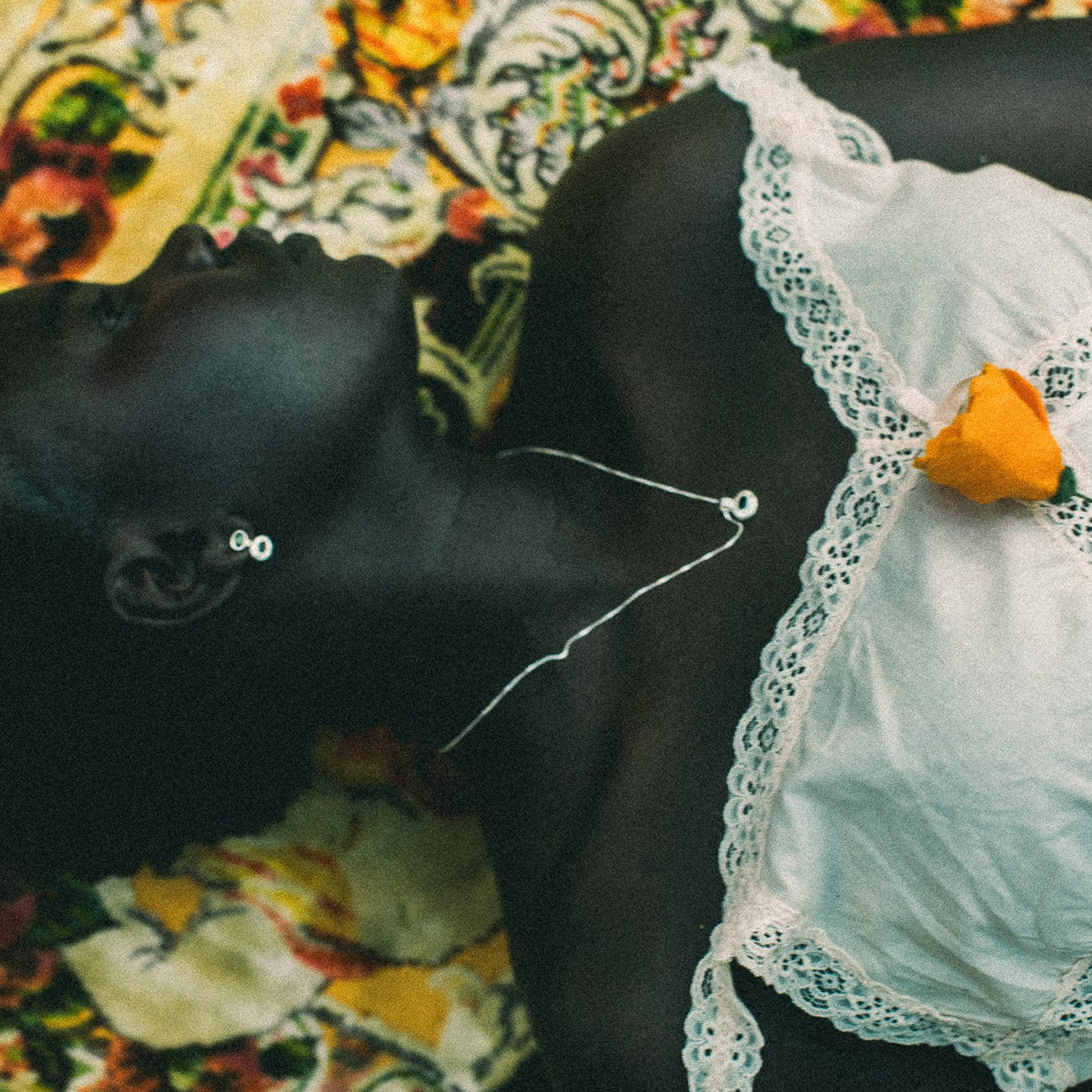 A close-up lifestyle shot of a person lying down wearing a white lace-trimmed top, with a small orange flower pinned to the chest, against a colorful patterned textile background.